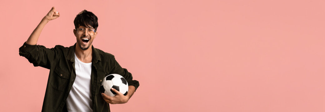 A young man with glasses is joyfully raising his arm in celebration while holding a soccer ball. He is smiling and dressed in casual clothing. The background is a solid pink color.