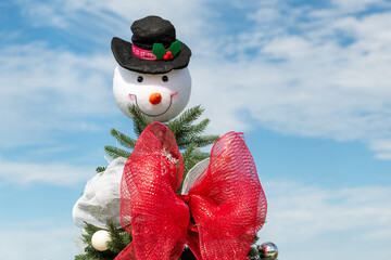 A white, round plush snowman face, smiling and wearing a black top hat as a Christmas tree topper. The decoration is colorful with a large red and white shimmery bow on the green fir tree outdoors.