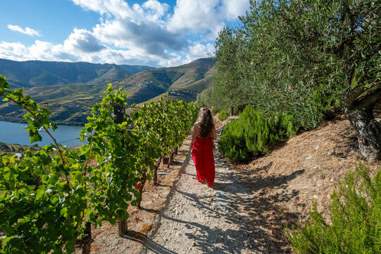 Young woman in red dress on walking path above terraced vineyards of Douro Valley wine region in Portugal under autumn cloudscape.
