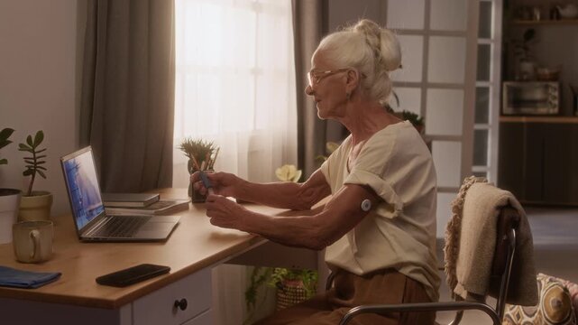 Medium shot of senior Caucasian woman sitting at table in living room, having telemedicine consultation with doctor on laptop, demonstrating diabetes patch and insulin pen while controlling disease