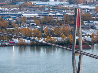 Naklejka premium Suspension Bridge Over River With Train Crossing Amid Industrial Complex and Autumn Trees