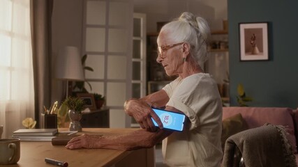 Medium shot of retired Caucasian woman with gray hair, living with autoimmune diabetes, sitting at home, placing smartphone to glucose monitor patch on arm, then looking at data on app screen - Powered by Adobe