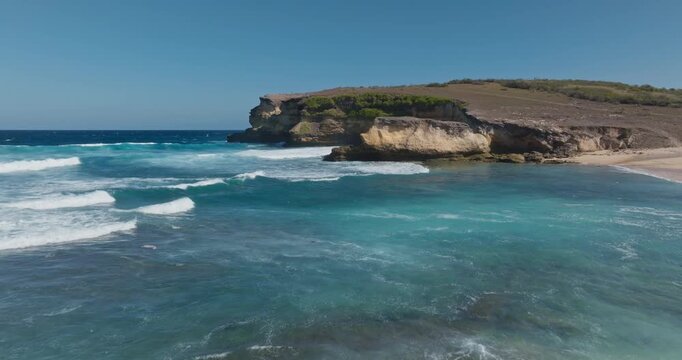 Penisok, Lombok, Indonesia: Ocean waves breaking on the sandy beach with rugged coastline cliff meeting the blue sky, creating a serene and natural tropical landscape. Wild nature. Aerial drone fligh