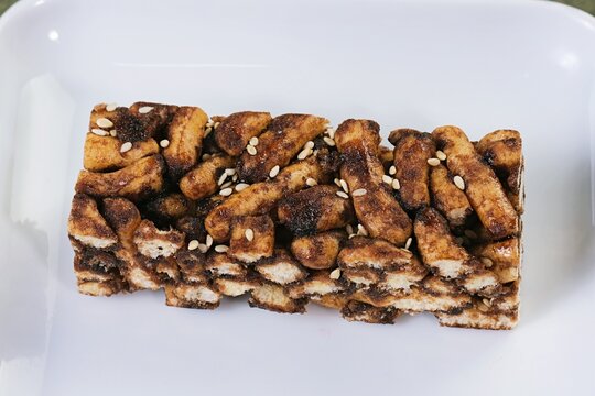 A dark brown sachima block presented on a clean white plate with soft lighting, focusing on crunchy caramel texture.