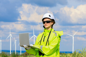Engineer woman in hardhat with laptop and blueprint working on wind turbines at electricity power station background. Renewable energy, wind turbine generate electricity to produce energy 