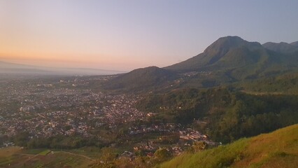 Obraz premium Sunset view of Batu City from Paralayang hill, showing a vibrant urban valley beneath a green mountain with warm sky hues and peaceful natural surroundings.