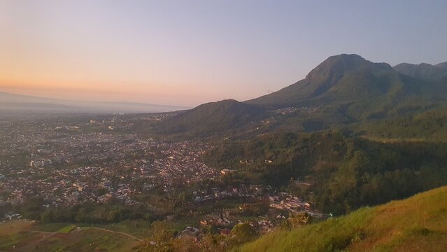 Sunset view of Batu City from Paralayang hill, showing a vibrant urban valley beneath a green mountain with warm sky hues and peaceful natural surroundings.