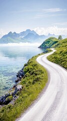 A winding gravel road curves along a rocky coastline beside a clear, calm ocean, leading towards distant, mist-shrouded mountains.