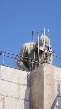 Concrete work, Construction worker wearing a hoodie and face covering, working on an outdoor concrete structure at height. Clear sky in the background
