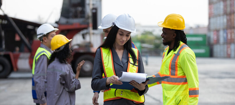 Female logistics supervisor in safety vest and helmet holding documents while inspecting operations at container shipping yard, representing leadership, planning and professional management.