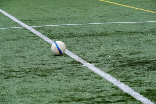 A white soccer ball with blue and red stripes rests on green synthetic turf near intersecting white and yellow field lines, capturing a moment of calm before play.