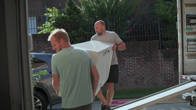 men move cabinet up ramp, pair of men carefully carry big cabinet into building during day, two men in white shirts transfer large cabinet into residential rowhouse with precise coordination