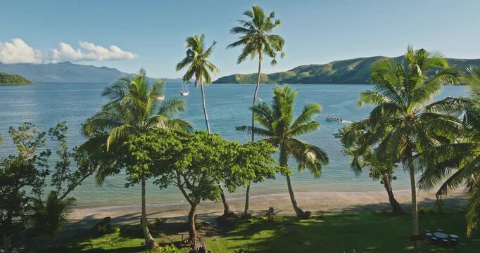 Fiji island beach tranquil tropical landscape with palm trees on sandy shore, expansive blue ocean and distant mountains under a clear blue sky. Aerial view drone flight over exotic nature paradise