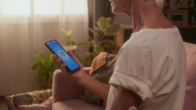 Medium side shot of elderly 70-year-old woman in white short sleeved shirt, with diabetes monitoring sensor patch, looking at blood sugar tracking app on smartphone, low number indicating hypoglycemia