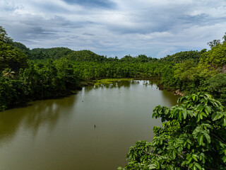 A calm freshwater lake is surrounded by dense tropical forest and hills under a cloudy sky. Siargao, Philippines.
