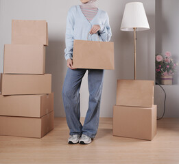 A young woman's body in a knitted sweater and jeans carries a jumbo paper shopping bag among a pile of cardboard boxes.