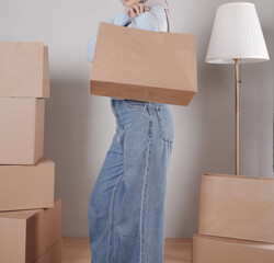 A young woman's body in a knitted sweater and jeans carries a jumbo paper shopping bag among a pile of cardboard boxes.