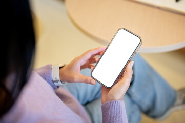 Woman in jeans holding white screen smartphone reading or watching as sitting indoors at cafe table.
