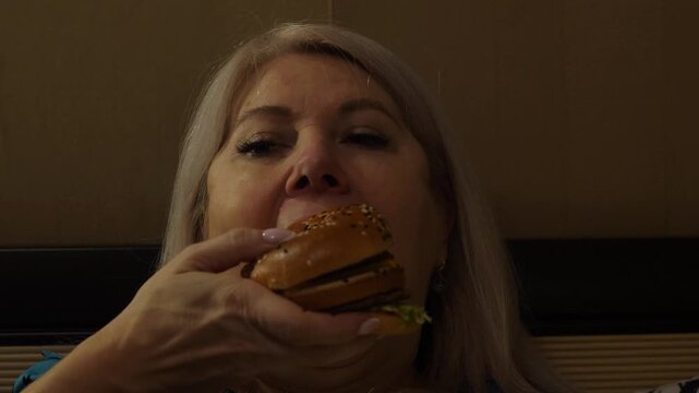 portrait of a middle-aged Caucasian woman eating a large appetizing hamburger while lying in bed in the evening, overeating problem, woman eats burger at nigh
