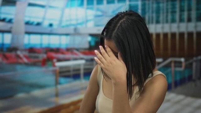 Woman indoors on cruise ship with sea view feels seasick while covering face with hand in casual tank top showing expressions of discomfort on modern vessel.