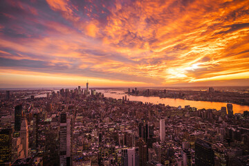 Obraz premium Aerial view of New York City at sunset with vibrant orange sky reflecting in the Hudson River and illuminated skyscrapers under dramatic clouds