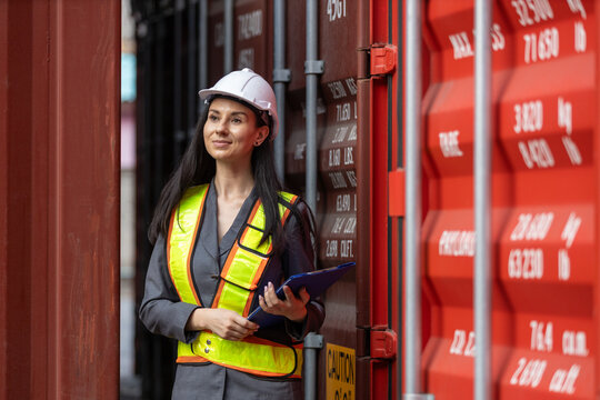 Female inspector in safety vest and helmet checking shipping container data for logistics and transportation industry, ensuring import-export accuracy and freight security.