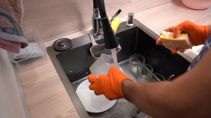 A person in orange gloves washes dishes in a modern sink, demonstrating effective cleaning techniques while using soap and a sponge to maintain kitchen hygiene..