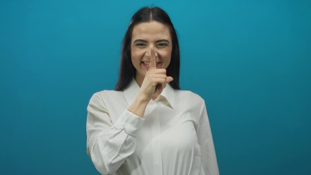 Young woman gesturing silence with finger on lips against blue wall background wearing white shirt showing playful attitude