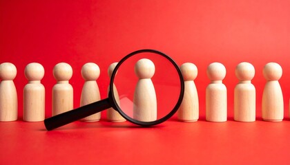 Magnifying glass focusing on wooden figures representing candidate selection on plain red background.