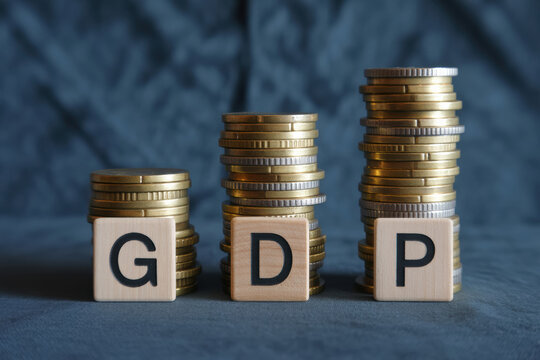 Three wooden scrabble tiles spelling gdp are placed in front of ascending stacks of gold and silver coins