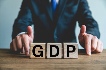 Businessman in a dark suit with a blue tie giving thumbs up while seated behind three wooden blocks spelling out GDP