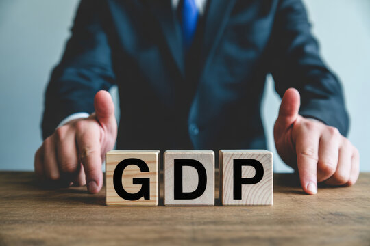 Businessman in a dark suit with a blue tie giving thumbs up while seated behind three wooden blocks spelling out GDP