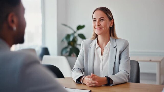白い壁と自然光に包まれたミニマルなオフィスで、白人女性が木のテーブル越しに同僚と穏やかに話しています。白いブラウスとライトグレーのジャケットが清潔感と信頼感を演出し、観葉植物が空間に柔らかいアクセントを加えます。