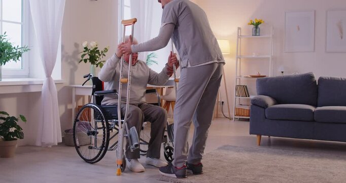 Young male caregiver or nurse helping senior patient with disability to get off the wheelchair using crutches. Physiotherapist or health care worker helping old disabled man in rehab. 4k video. - Powered by Adobe