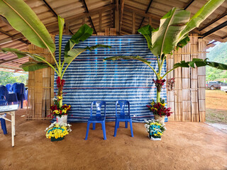 A minimalist wedding stage of the Lahu hill tribe in northern Thailand, featuring banana trees, flowers, and blue chairs for a traditional countryside ceremony.