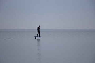 A young unknown man glides smoothly over the calm sea morning on an electric hydrofoil surfboard,...