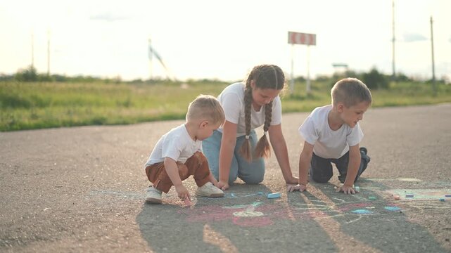 A girl, a boy, and a toddler sit on the street, the kids using chalk to draw colorful shapes. The child in brown pants leans forward, the girl with braids smiles, and the boy focuses. Outdoor play