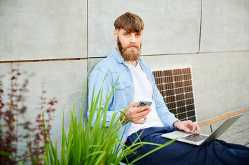 Bearded man works on laptop on lap and smartphone. Solar panel nearby, emphasizing sustainable, modern eco-friendly workspace that combines technology and sustainability.
