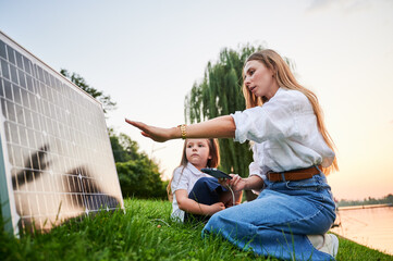 Mother connecting smartphone to photovoltaic solar panel for charging, explains its workings to daughter, sit on grass by lake. Integration of sustainable renewable energy into everyday life.
