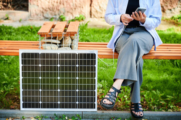 Woman using smartphone connected to solar panel. Cropped view. Integration of sustainable renewable energy into everyday life, demonstrating practical use of solar power for charging devices.