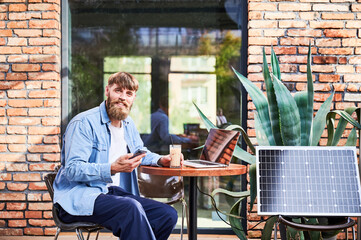 Man works on laptop and smartphone at outdoor table, with glass of iced coffee. Solar panel nearby, emphasizing sustainable, modern eco-friendly workspace that combines technology and sustainability.