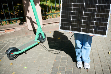 Woman using portable solar panel to charge scooter, highlighting integration of sustainable renewable energy. Green electric scooter connected to solar panel, emphasizing eco-friendly transportation.