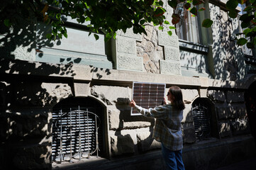 Woman holding photovoltaic solar panel in front of historical building, wearing plaid shirt and sunglasses. Concept of integration of sustainable renewable energy sources into old architecture.