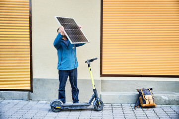 Male person holds photovoltaic solar panel, for charging electric scooter in urban settings. Integration of solar power as sustainable energy source, promoting eco-friendly urban transportation.