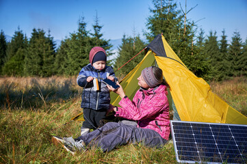 Young child and mother play with smartphone while charging with photovoltaic solar panel near tourist tent in summer. Integration of renewable energy in outdoor camping activities.