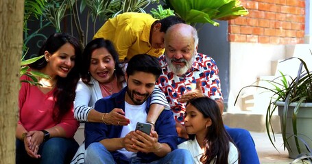 Indian family of six using smartphone sitting on steps outdoors in home garden, surfing internet or checking social media together, cheerful multi-generation portrait using technology - Powered by Adobe