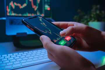 Close-up of a trader hands analyzing financial charts or cryptocurrency prices on a smartphone and laptop trading app, showing buy and sell buttons during online stock buying or crypto investing.
