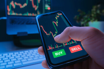 Close up of a trader hand holds a smartphone with a stock market trading app on the screen, buying cryptocurrency and investing in stocks at their desk with laptop. Copy space