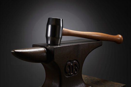 classic blacksmith's cross-peen hammer resting on top of a heavy, worn, black iron anvil against a dark studio background