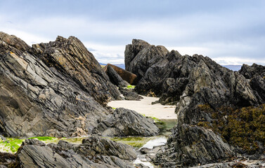 Rugged coastal rocks with a small sandy cove on the Isle of Iona, Scotland. Natural seaside landscape with dramatic rock formations and cloudy sky.